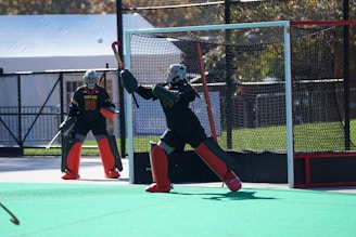 Two field hockey goalkeepers are actively engaged on a pitch, wearing protective gear including helmets, pads, and gloves. One goalkeeper is in a ready stance, poised to block the ball, while the other stands nearby. The vibrant green surface contrasts with their dark uniforms.