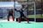 Two field hockey goalkeepers are actively engaged on a pitch, wearing protective gear including helmets, pads, and gloves. One goalkeeper is in a ready stance, poised to block the ball, while the other stands nearby. The vibrant green surface contrasts with their dark uniforms.