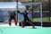 Two field hockey goalkeepers are actively engaged on a pitch, wearing protective gear including helmets, pads, and gloves. One goalkeeper is in a ready stance, poised to block the ball, while the other stands nearby. The vibrant green surface contrasts with their dark uniforms.