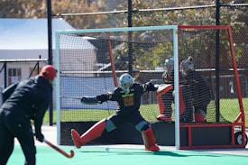 A field hockey game is taking place with a player in black and red gear attempting to score against a goalie. The goalie is wearing protective gear, including a helmet and red leg pads, standing in front of the goal. The netted goal is positioned on a synthetic green field. Other players in protective helmets are visible near the goal.