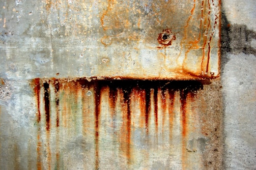 A weathered concrete wall with prominent rust stains running vertically across its surface, suggesting corrosion and decay. The texture of the concrete is rough, with visible aggregate and discoloration.