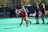A group of athletes wearing red uniforms are on a green field, practicing field hockey. One player is in the foreground actively swinging a hockey stick towards a white ball. The background features additional players and a row of benches with water bottles, and there are trees beyond the field adding a touch of nature to the setting.