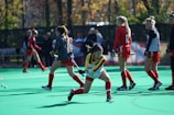 A group of female field hockey players are actively engaged in a practice session on a green turf field. One player is in a dynamic position, preparing to strike a ball with her stick, while others in red and black uniforms are visible in the background, some with numbered jerseys.