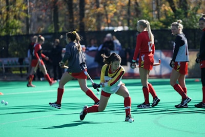 A group of female field hockey players are actively engaged in a practice session on a green turf field. One player is in a dynamic position, preparing to strike a ball with her stick, while others in red and black uniforms are visible in the background, some with numbered jerseys.