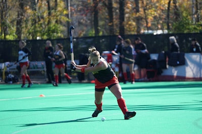 A field hockey player is in mid-swing, about to hit the ball on a green artificial turf. The player is wearing a black jersey and red shorts with matching socks. In the background, several other players and people, possibly a coach or spectators, are visible near the sidelines.
