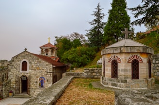 A stone chapel with a red-tiled roof and a golden cross is situated on the left, surrounded by lush greenery. Adjacent to it on the right is a circular stone structure with decorative archways and a cross on top. The background is a landscape filled with trees and shrubs.