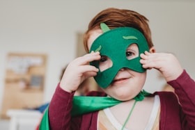 Um menino ruivo segurando uma máscara de super herói verde em seu rosto