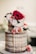 A two-tier semi-naked cake decorated with fresh flowers, including vibrant red, pink, and white blooms with green leaves. Caramel drizzle elegantly cascades down the sides of the cake, creating a rustic yet elegant look. The cake sits on a simple white stand in a softly blurred background setting.