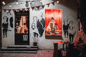 A cozy bookstore with a warmly lit window display featuring red curtains and books stacked inside. The exterior walls are adorned with large, artistic black-and-white murals of people. String lights hang above, contributing to the inviting atmosphere, and there's a small round table with chairs outside.