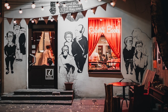 A cozy bookstore with a warmly lit window display featuring red curtains and books stacked inside. The exterior walls are adorned with large, artistic black-and-white murals of people. String lights hang above, contributing to the inviting atmosphere, and there's a small round table with chairs outside.