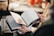 A person is holding an open book with handwritten notes, flipping through the pages. The background is blurred, giving the impression of an indoor setting with soft lighting.