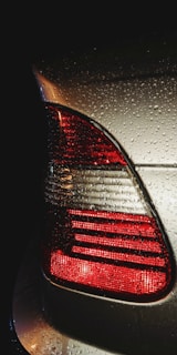 The image captures the rear end of a car with its taillight illuminated. Water droplets are visible on the surface, indicating recent rain or a wash. The scene appears to be set in low light, highlighting the red and silver colors.