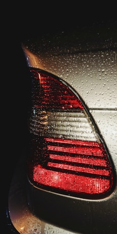 The image captures the rear end of a car with its taillight illuminated. Water droplets are visible on the surface, indicating recent rain or a wash. The scene appears to be set in low light, highlighting the red and silver colors.