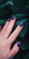 A hand with well-manicured nails featuring a dark blue and pink artistic pattern is placed against a backdrop of dark green leaves. The nails have a glossy finish and the hand wears a silver ring.