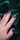 A hand with well-manicured nails featuring a dark blue and pink artistic pattern is placed against a backdrop of dark green leaves. The nails have a glossy finish and the hand wears a silver ring.