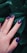 A hand with well-manicured nails featuring a dark blue and pink artistic pattern is placed against a backdrop of dark green leaves. The nails have a glossy finish and the hand wears a silver ring.