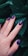 A hand with well-manicured nails featuring a dark blue and pink artistic pattern is placed against a backdrop of dark green leaves. The nails have a glossy finish and the hand wears a silver ring.