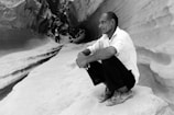 A man squats on a rocky surface within a canyon or narrow gorge, with intricate stone walls surrounding him. He appears contemplative, dressed in a white shirt and dark pants. Another person can be seen in the distance, walking through the winding path of the canyon.