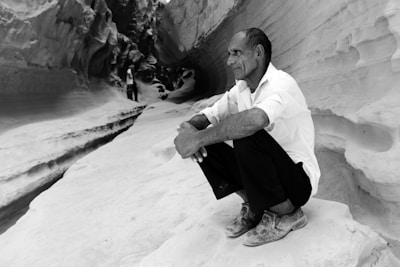 A man squats on a rocky surface within a canyon or narrow gorge, with intricate stone walls surrounding him. He appears contemplative, dressed in a white shirt and dark pants. Another person can be seen in the distance, walking through the winding path of the canyon.