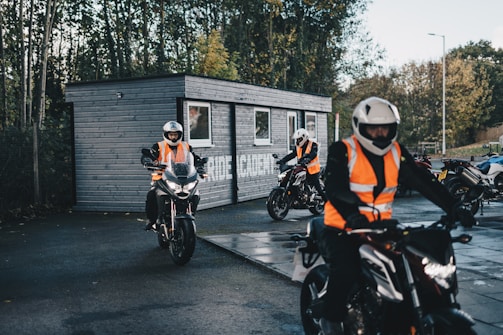 Several motorcyclists wearing bright orange safety vests and helmets are riding around a small, dark grey wooden building, likely used for training or as a course base. The scene is outdoors with a background of trees and a clear sky, suggesting a well-lit day. The riders appear to be practicing or attending a training session.