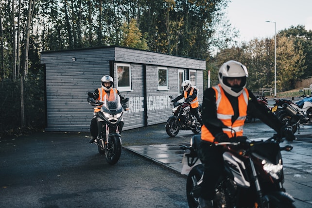 Several motorcyclists wearing bright orange safety vests and helmets are riding around a small, dark grey wooden building, likely used for training or as a course base. The scene is outdoors with a background of trees and a clear sky, suggesting a well-lit day. The riders appear to be practicing or attending a training session.