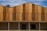 A modern building facade featuring vertical wooden panels of varying tones, complemented by horizontal louvered sections that possibly serve as ventilation. The lower part consists of a textured stone wall, contrasting with the smooth wood. The roofline has an angular, stepped design.