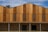 A modern building facade featuring vertical wooden panels of varying tones, complemented by horizontal louvered sections that possibly serve as ventilation. The lower part consists of a textured stone wall, contrasting with the smooth wood. The roofline has an angular, stepped design.