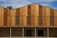 A modern building facade featuring vertical wooden panels of varying tones, complemented by horizontal louvered sections that possibly serve as ventilation. The lower part consists of a textured stone wall, contrasting with the smooth wood. The roofline has an angular, stepped design.