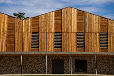 A modern building facade featuring vertical wooden panels of varying tones, complemented by horizontal louvered sections that possibly serve as ventilation. The lower part consists of a textured stone wall, contrasting with the smooth wood. The roofline has an angular, stepped design.