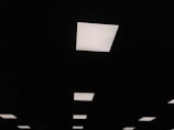A series of rectangular ceiling lights are illuminated against a dark ceiling, creating a pattern of bright white shapes on a black background.