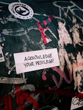 A sticker with the text 'ACKNOWLEDGE YOUR PRIVILEGE!' is placed on a wall covered with various graffiti and stickers. The wall is heavily marked with different colors and patterns, giving it a chaotic and vibrant look.