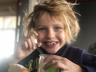 A happy child reaching for a spoonful of supplement syrup with a soft green backdrop.