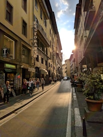 A narrow street flanked by tall, historic buildings with a 'Ristorante' sign. Pedestrians are casually walking along the sidewalk and window shopping. The atmosphere is warm, highlighted by the setting sun casting a golden glow on the buildings.