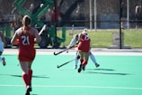 Several field hockey players are actively engaged in a game on a bright green turf field. Two players are wearing red uniforms with numbers visible on their backs, while a player in a white uniform is seen attempting to maneuver past them. The background features a fence and some out-of-focus spectators or equipment.