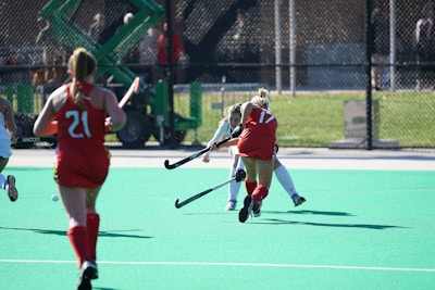 Several field hockey players are actively engaged in a game on a bright green turf field. Two players are wearing red uniforms with numbers visible on their backs, while a player in a white uniform is seen attempting to maneuver past them. The background features a fence and some out-of-focus spectators or equipment.