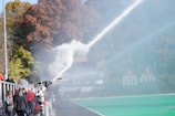 A camera is positioned by the sidelines of a sports field capturing a spray of water. There are people in casual clothing walking along a path beside a green field. In the background, there are trees with autumnal foliage.