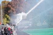 A camera is positioned by the sidelines of a sports field capturing a spray of water. There are people in casual clothing walking along a path beside a green field. In the background, there are trees with autumnal foliage.