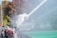 A camera is positioned by the sidelines of a sports field capturing a spray of water. There are people in casual clothing walking along a path beside a green field. In the background, there are trees with autumnal foliage.