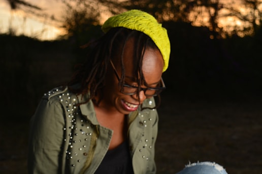 A person with glasses is laughing joyfully outdoors during a sunset. They are wearing an olive green jacket adorned with studs and a bright yellow head wrap.