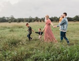 A family of four is walking through a lush green meadow. The woman is wearing a red floral dress and a hat, while holding hands with two young boys. The man is wearing a denim jacket and is holding a baby in his arms. String lights are visible in the background, adding a whimsical touch to the setting under a cloudy sky.