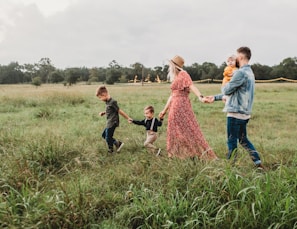 A family of four is walking through a lush green meadow. The woman is wearing a red floral dress and a hat, while holding hands with two young boys. The man is wearing a denim jacket and is holding a baby in his arms. String lights are visible in the background, adding a whimsical touch to the setting under a cloudy sky.