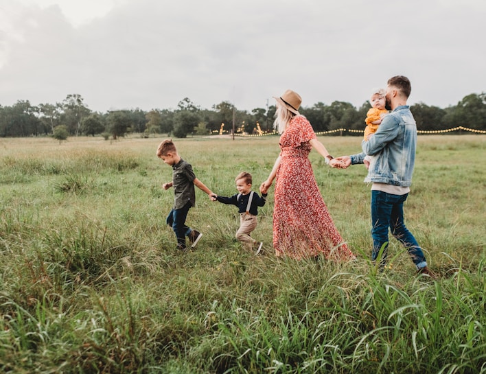 A family of four is walking through a lush green meadow. The woman is wearing a red floral dress and a hat, while holding hands with two young boys. The man is wearing a denim jacket and is holding a baby in his arms. String lights are visible in the background, adding a whimsical touch to the setting under a cloudy sky.