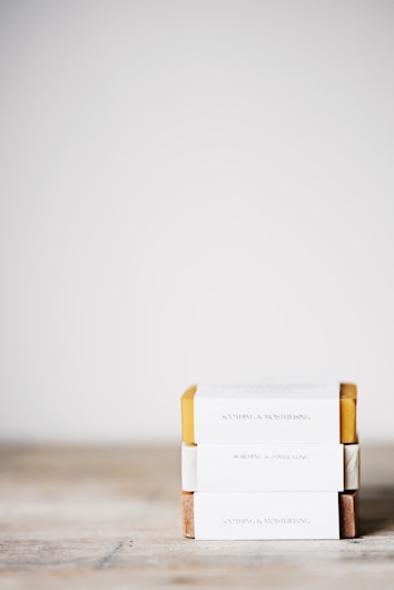 Three neatly stacked rectangular soap bars with minimalistic packaging, each wrapped with a paper label indicating qualities such as soothing and moisturizing. The stack is placed on a wooden surface against a plain, light-colored background.