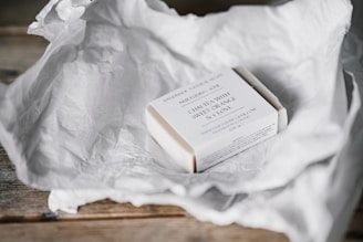 A bar of soap wrapped in a minimalistic white paper packaging rests on a crumpled sheet of white tissue paper, placed on a wooden surface. The packaging includes text that specifies the soap's scent as 'chai tea with sweet orange and clove.'