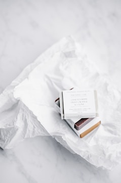 A small white box containing soap or a similar product is placed on crumpled white tissue paper atop a white marble surface. The box has text on it mentioning 'green tea with sweet orange & clove.' The packaging is minimalist and clean.