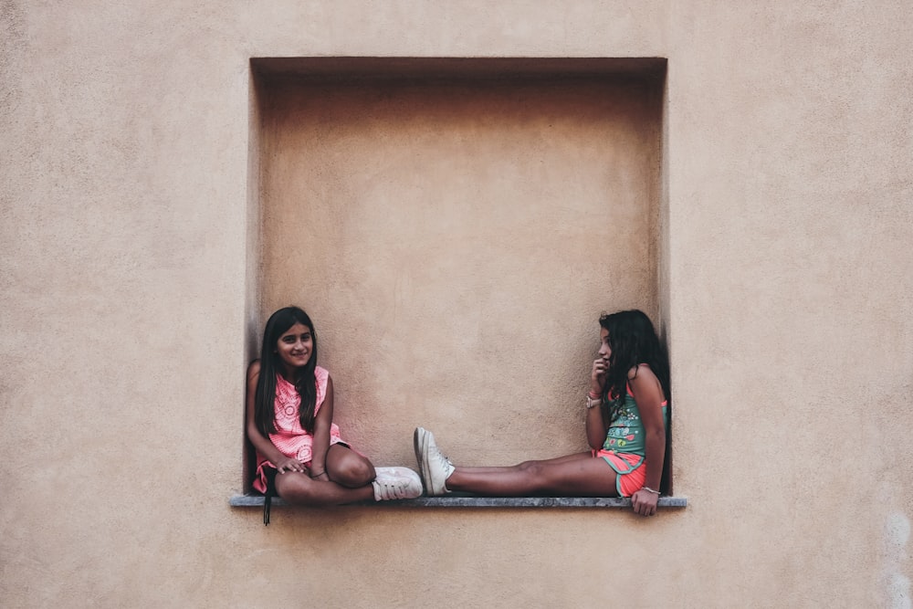 two girl in pink and green shirts sitting on wall shelf