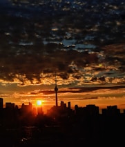 A skyline silhouette with a prominent tower is set against a vibrant sunset. The sky features dramatic clouds with hues of orange, yellow, and dark blue, adding depth to the scene. The sun is partially hidden behind the buildings, creating a warm glow.