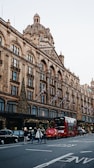 A busy street scene featuring an iconic department store adorned with festive lights and decorations. The architecture is elaborate with a large dome and multiple arched windows. Red double-decker buses and black cabs are present on the street, along with pedestrians crossing the road. Flags are displayed along the building facade.