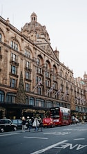 A busy street scene featuring an iconic department store adorned with festive lights and decorations. The architecture is elaborate with a large dome and multiple arched windows. Red double-decker buses and black cabs are present on the street, along with pedestrians crossing the road. Flags are displayed along the building facade.