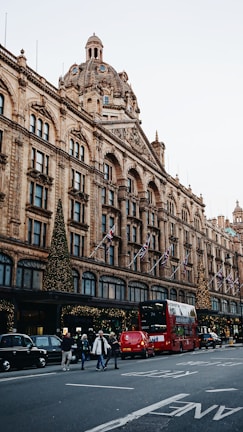 A busy street scene featuring an iconic department store adorned with festive lights and decorations. The architecture is elaborate with a large dome and multiple arched windows. Red double-decker buses and black cabs are present on the street, along with pedestrians crossing the road. Flags are displayed along the building facade.